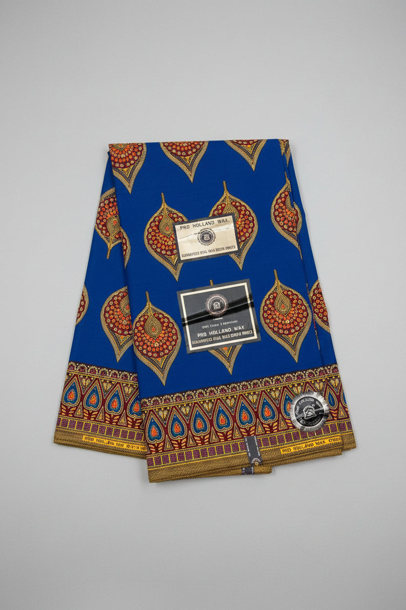 A folded blue African wax print fabric with gold, red, and orange traditional patterns, featuring two paper brand tags and a circular logo sticker, displayed on a textured gray background.