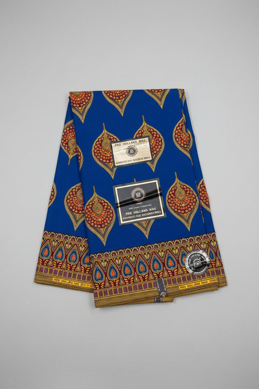 A folded blue African wax print fabric with gold, red, and orange traditional patterns, featuring two paper brand tags and a circular logo sticker, displayed on a textured gray background.